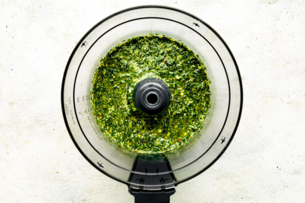 An overhead shot of blended chimichurri sauce in a food processor atop a white textured surface.