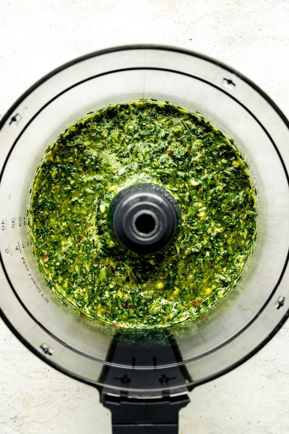 An overhead shot of blended chimichurri sauce in a food processor atop a white textured surface.