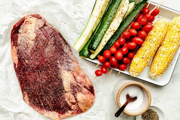 An overhead shot of prepared ingredients atop a white surface: seasoned flank steak, seasoned vegetables on a sheet pan, a jar of salt and bowl of pepper.