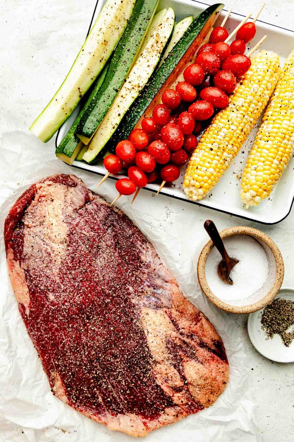 An overhead shot of prepared ingredients atop a white surface: seasoned flank steak, seasoned vegetables on a sheet pan, a jar of salt and bowl of pepper.