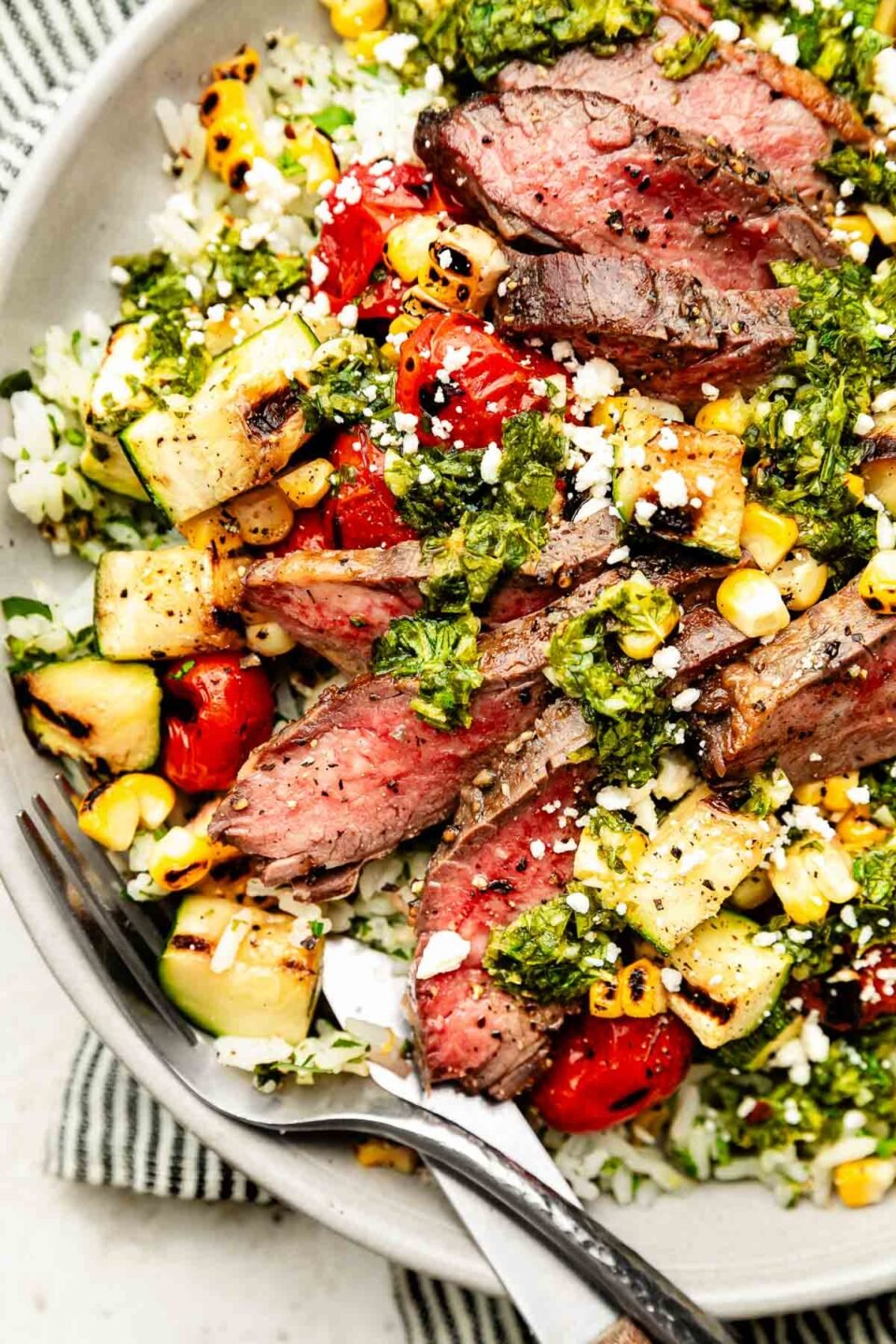 An overhead shot of a chimichurri steak bowl atop a striped cloth on a white textured surface.