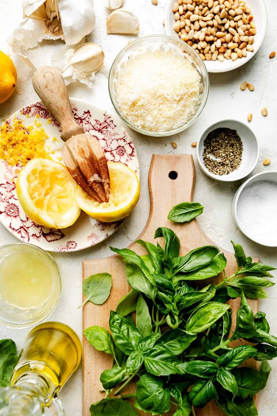 An overhead shot of ingredients displayed on a white textured surface on a cutting board and a variety of dishes: basil, parmesan, lemons, olive oil, pine nuts, garlic, salt and pepper.