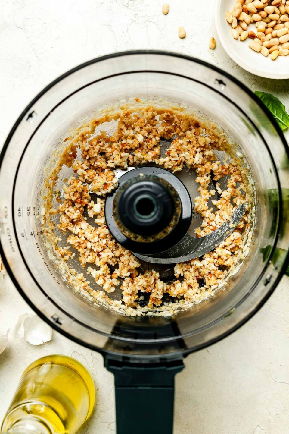 An overhead shot of a food processor with chopped pine nuts, sitting on a white surface. Olive oil and a bowl of pine nuts are sitting next to the food processor.