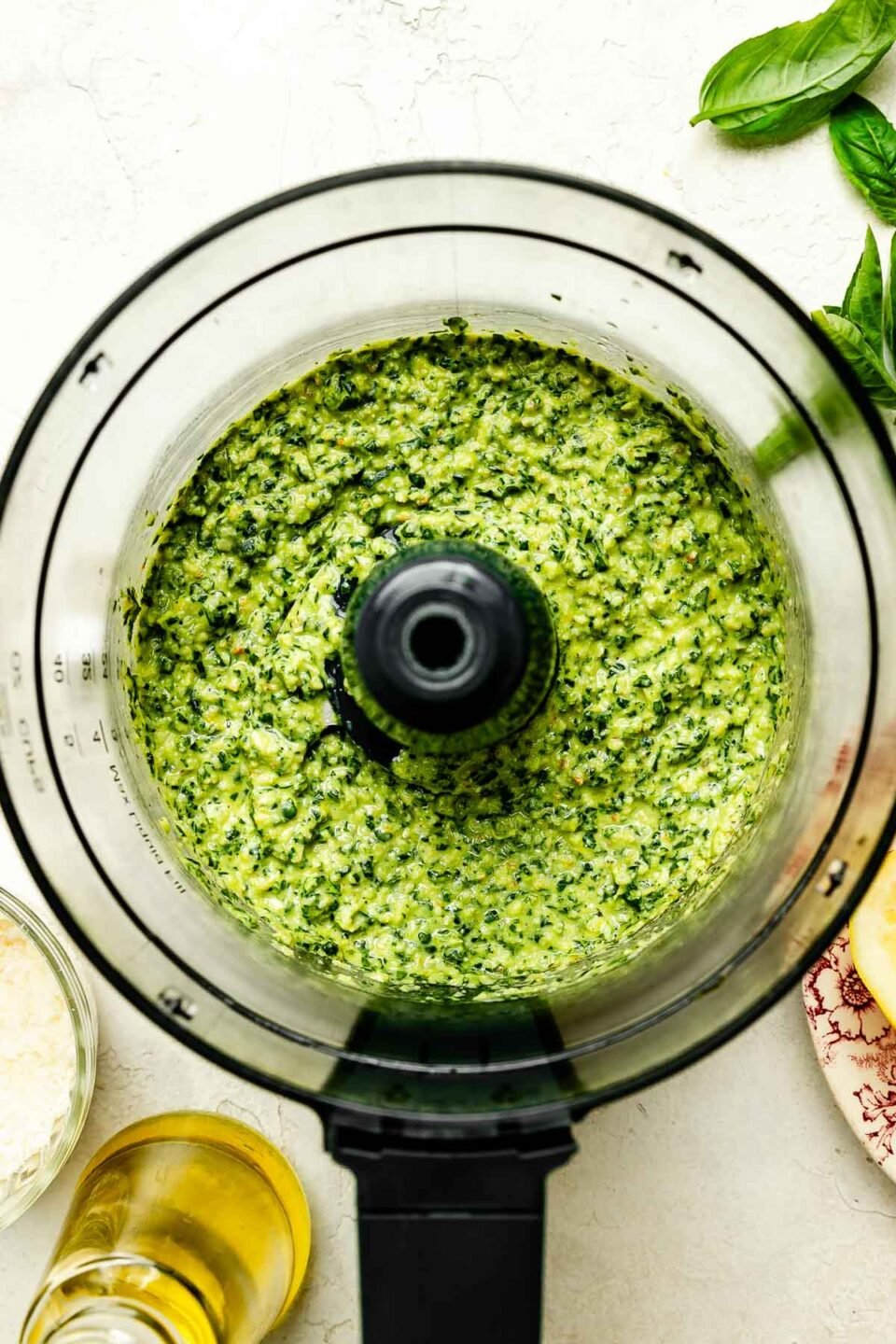 An overhead shot of a food processor full of lemon basil pesto atop a textured white surface. A bowl of grated parmesan, fresh basil leaves and olive oil sit alongside it.