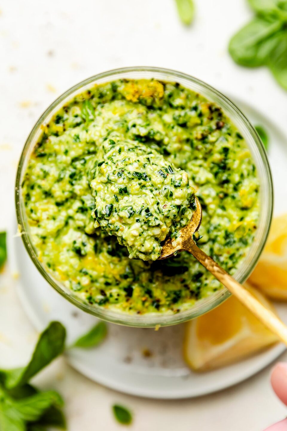 A close-up overhead shot of a spoon holding up basil pesto from a jar. The jar sits on a white plate with lemon wedges atop a white surface.