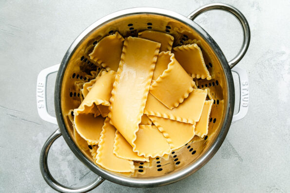 An overhead shot of lasagna noodles in a colander over a pot on top of a light grey surface.