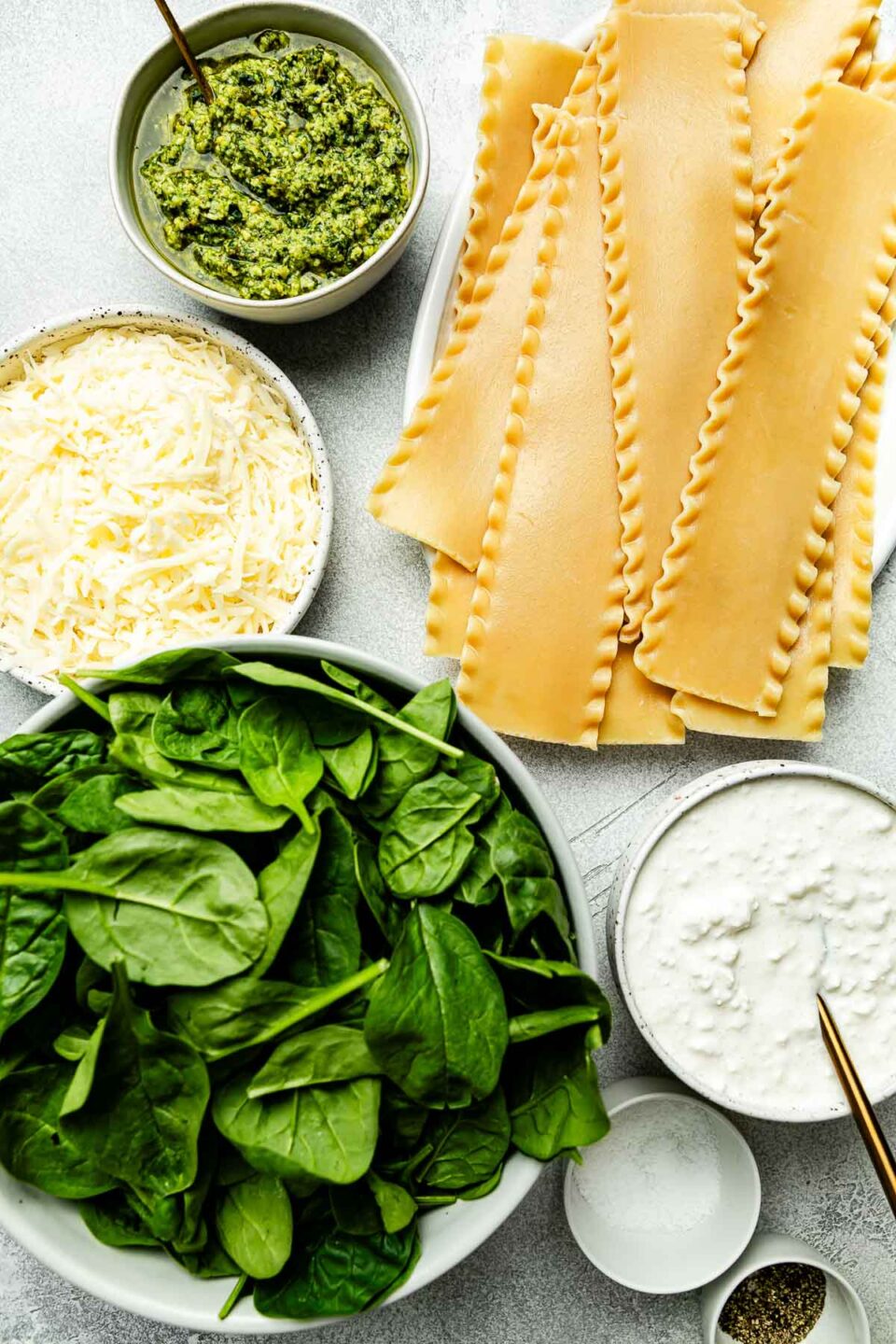 Ingredients are displayed on a light grey surface: lasagna noodles, basil pesto, spinach, cottage cheese, mozzarella, and salt and pepper.