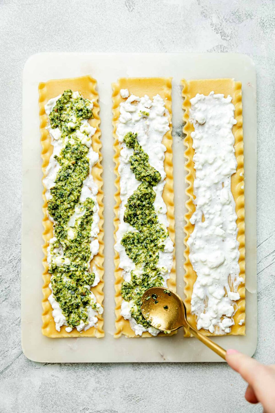 An overhead shot of a gold spoon spreading pesto over cottage cheese on lasagna noodles on a white board atop a light grey surface.