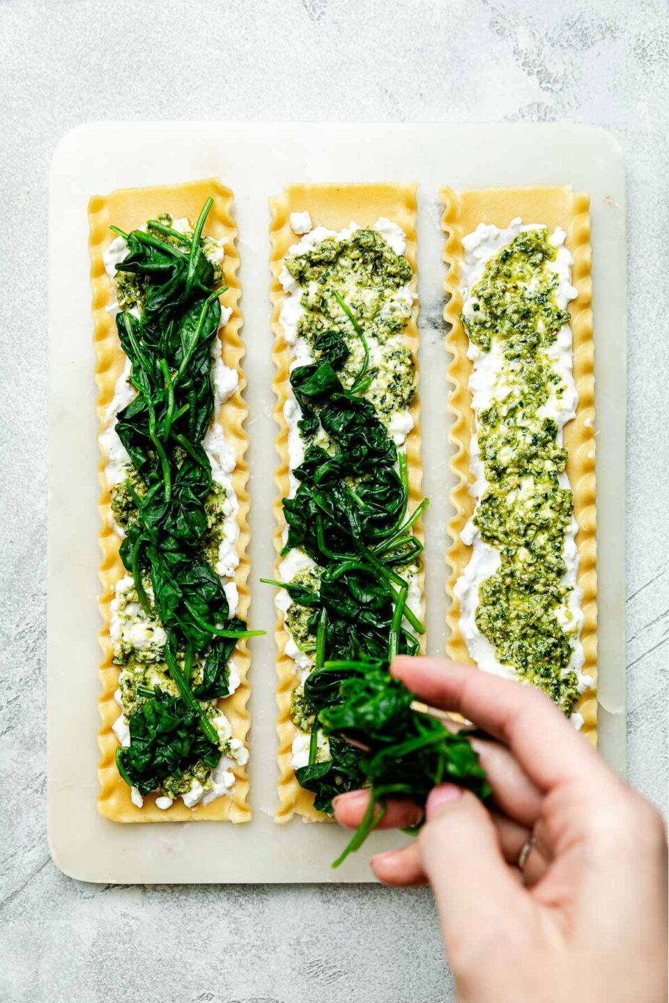 An overhead shot of a hand sprinkling spinach onto the cottage cheese and pesto lasagna noodles on a white board atop a light grey surface.