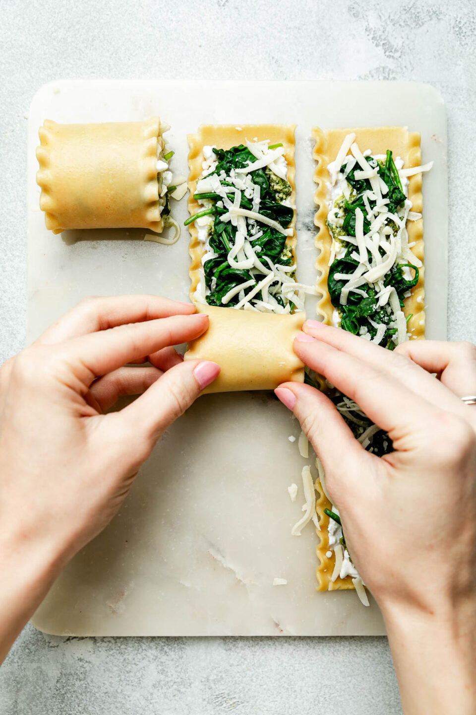 An overhead shot of a woman's hands rolling up lasagna rolls on a white board atop a light grey surface.