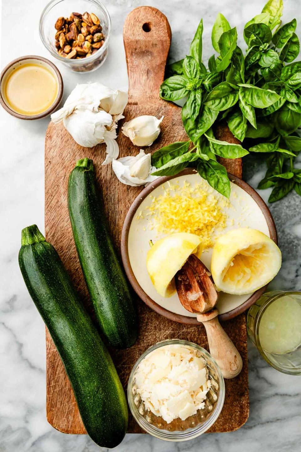 Ingredients displayed on a wooden cutting board and white marbled surface: Zucchini, lemons, basil, parmesan, garlic, pistachios and olive oil.