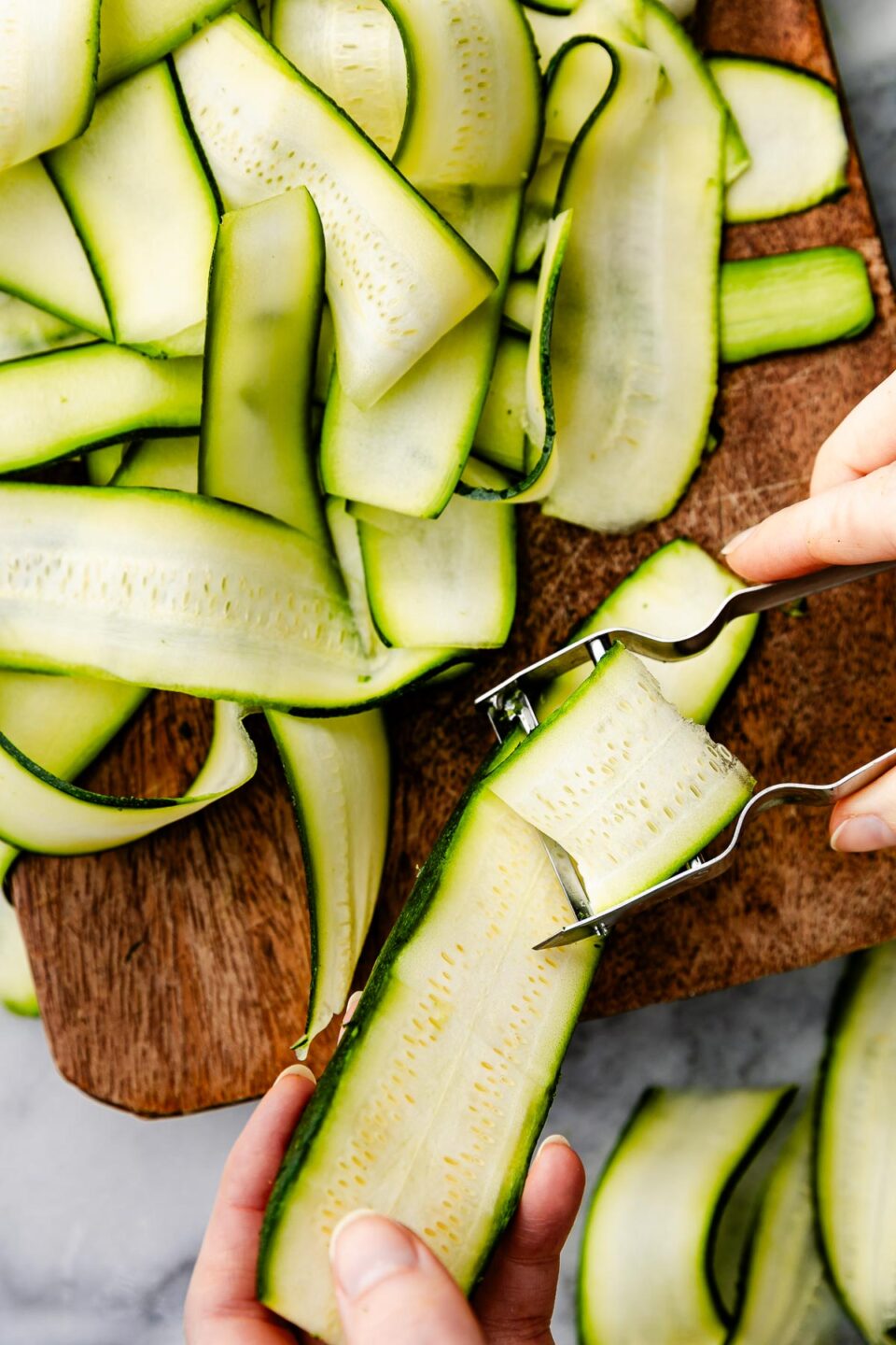 An overhead shot of zucchini being shaved into ribbons atop a wooden board.