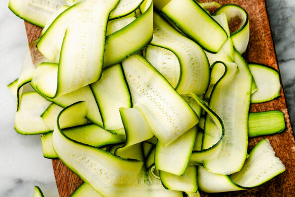 A close-up overhead shot of shaved zucchini ribbons displayed on a wooden board atop a white marbled surface.