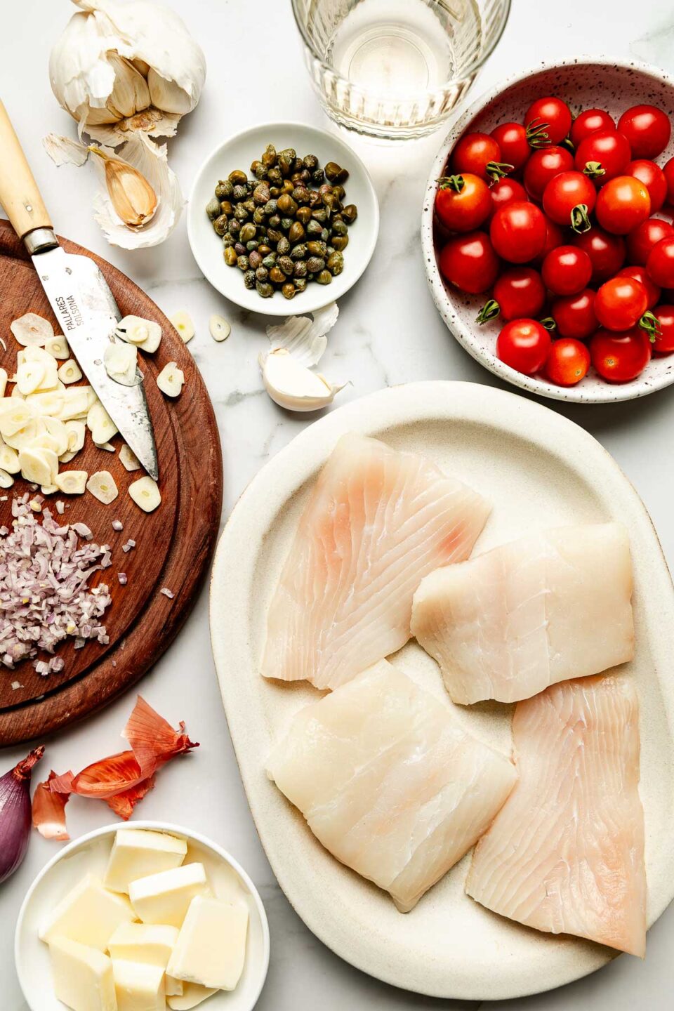Ingredients are displayed on a white marbled surface: a stoneware dish of raw cod filets, a bowl of cherry tomatoes, sliced butter, garlic, capers and shallot.