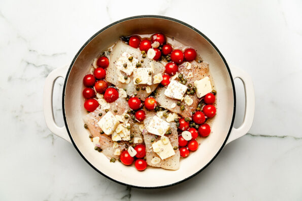An overhead shot of uncooked ingredients in a skillet atop a white marbled surface: fish filets, pats of butter, cherry tomatoes, capers, garlic and shallots.