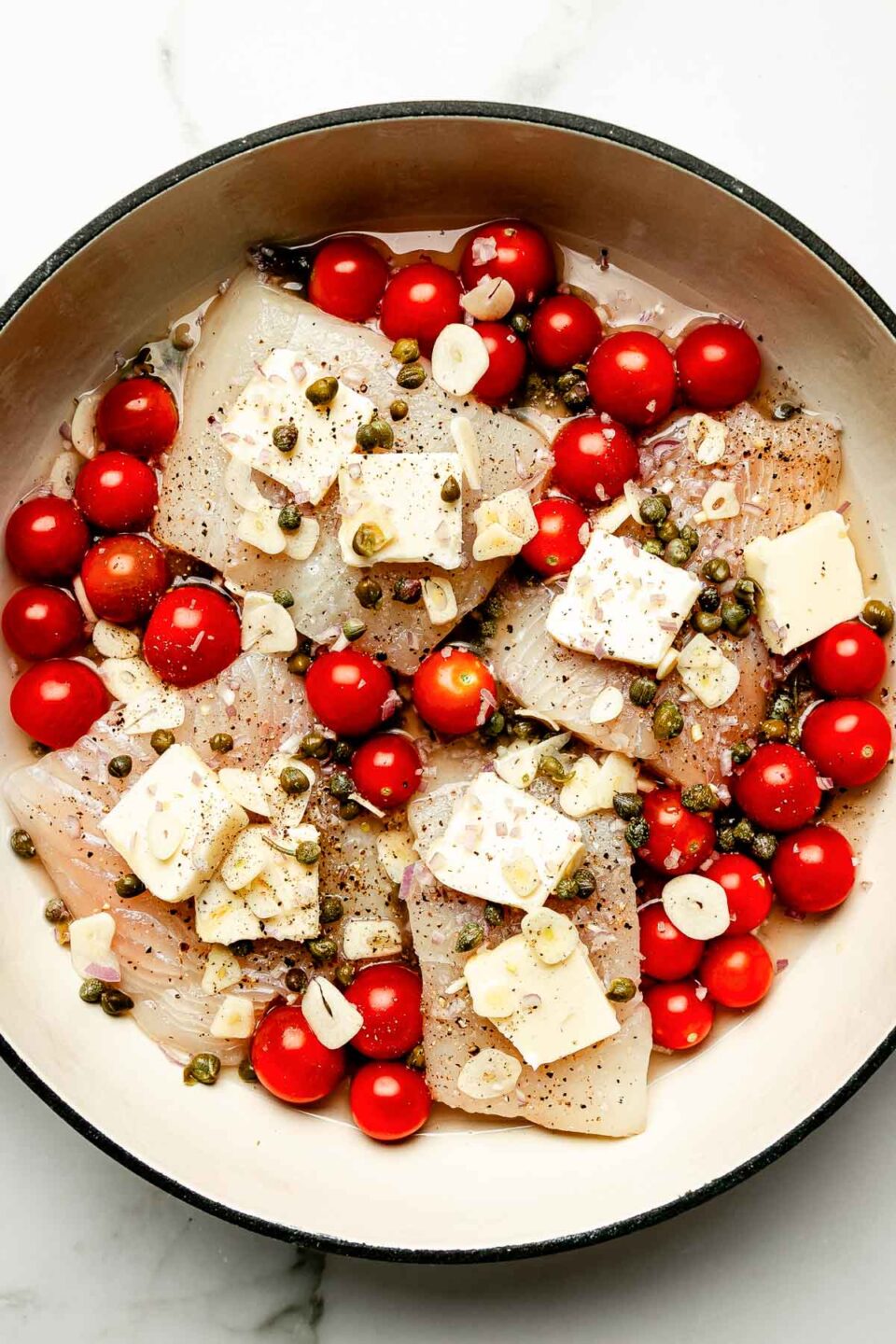 An overhead shot of uncooked ingredients in a skillet atop a white marbled surface: cod filets, pats of butter, cherry tomatoes, capers, garlic and shallots.