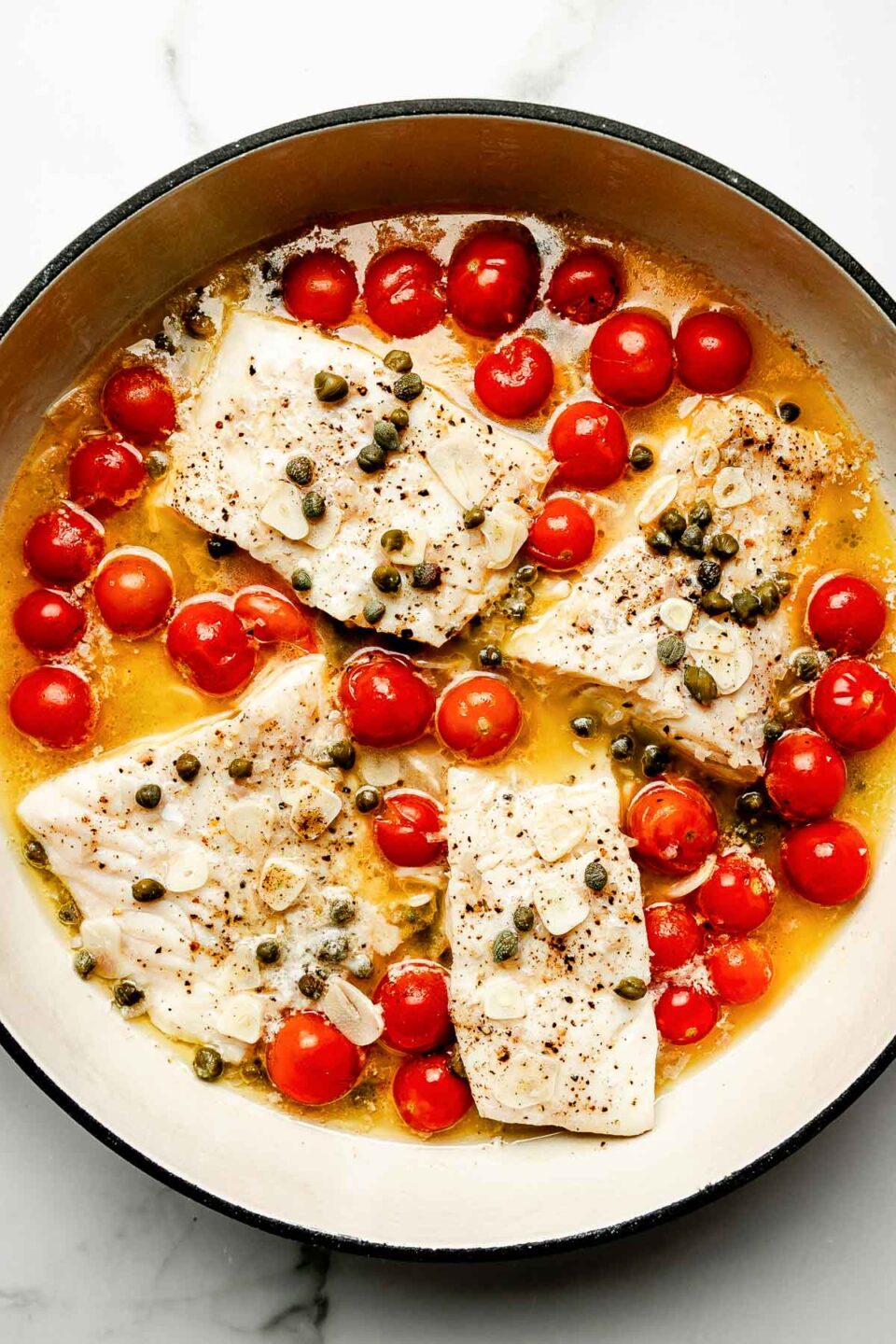 An overhead shot of steamed cod and tomatoes in white wine sauce in a white skillet atop a white marbled surface.