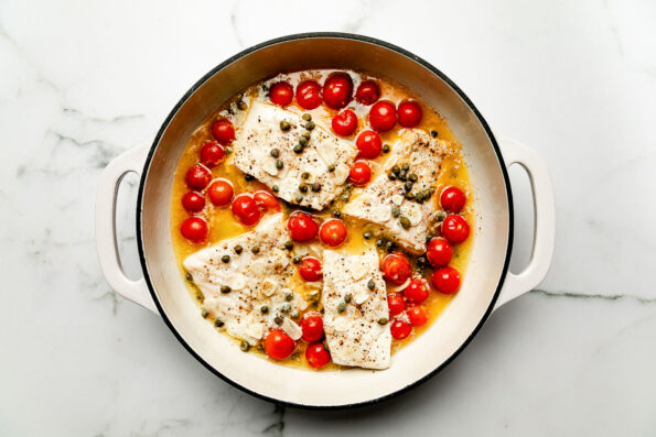 An overhead shot of steamed fish and tomatoes in white wine sauce in a white skillet atop a white marbled surface.