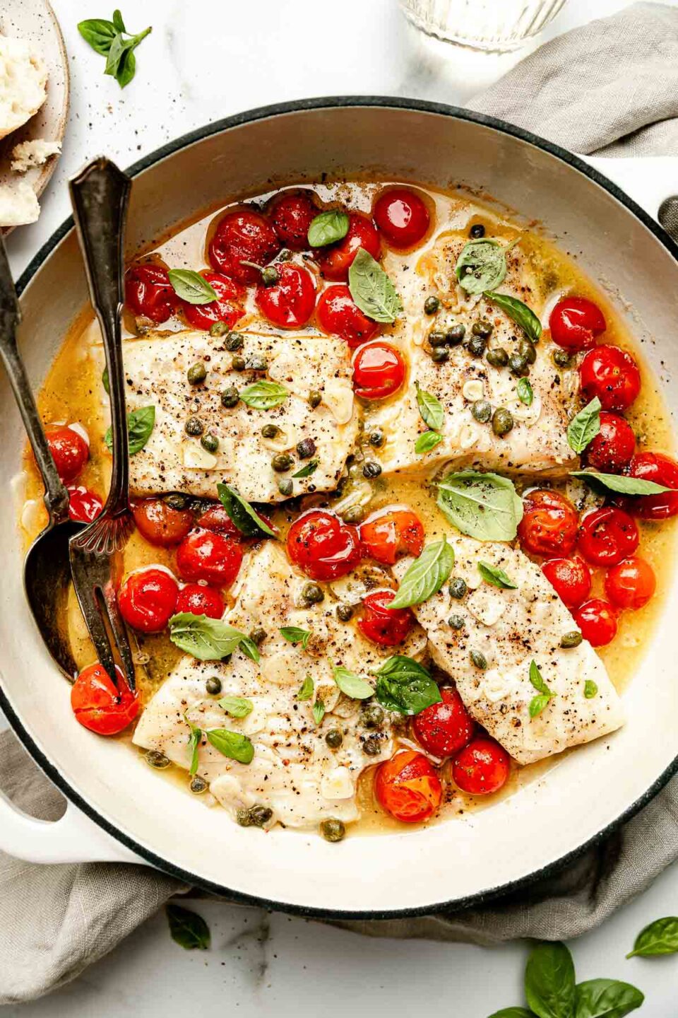 An overhead shot of cooked white wine steamed fish and cherry tomatoes in a white skillet atop a white surface. A beige cloth surrounds the skillet.