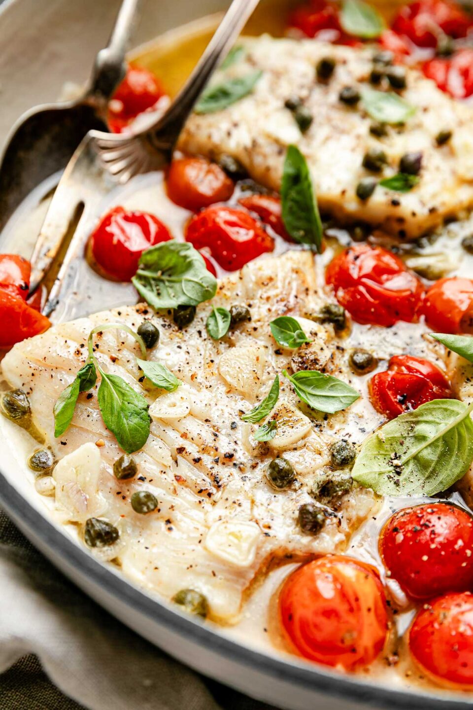A close-up macro shot of steamed fish topped with basil leaves, capers and garlic, and surrounded by steamed cherry tomatoes, in a white skillet.