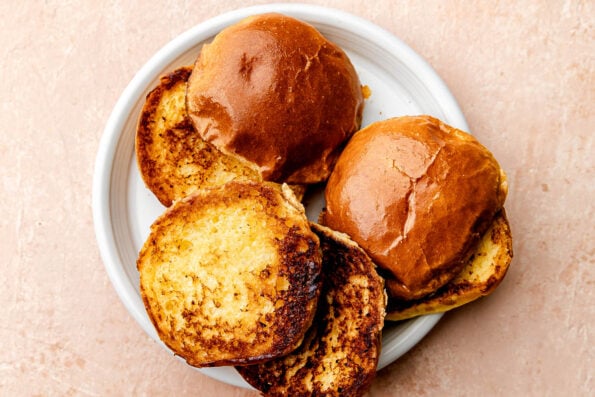 An overhead shot of toasted burger buns on a white plate sitting atop a light pink surface.