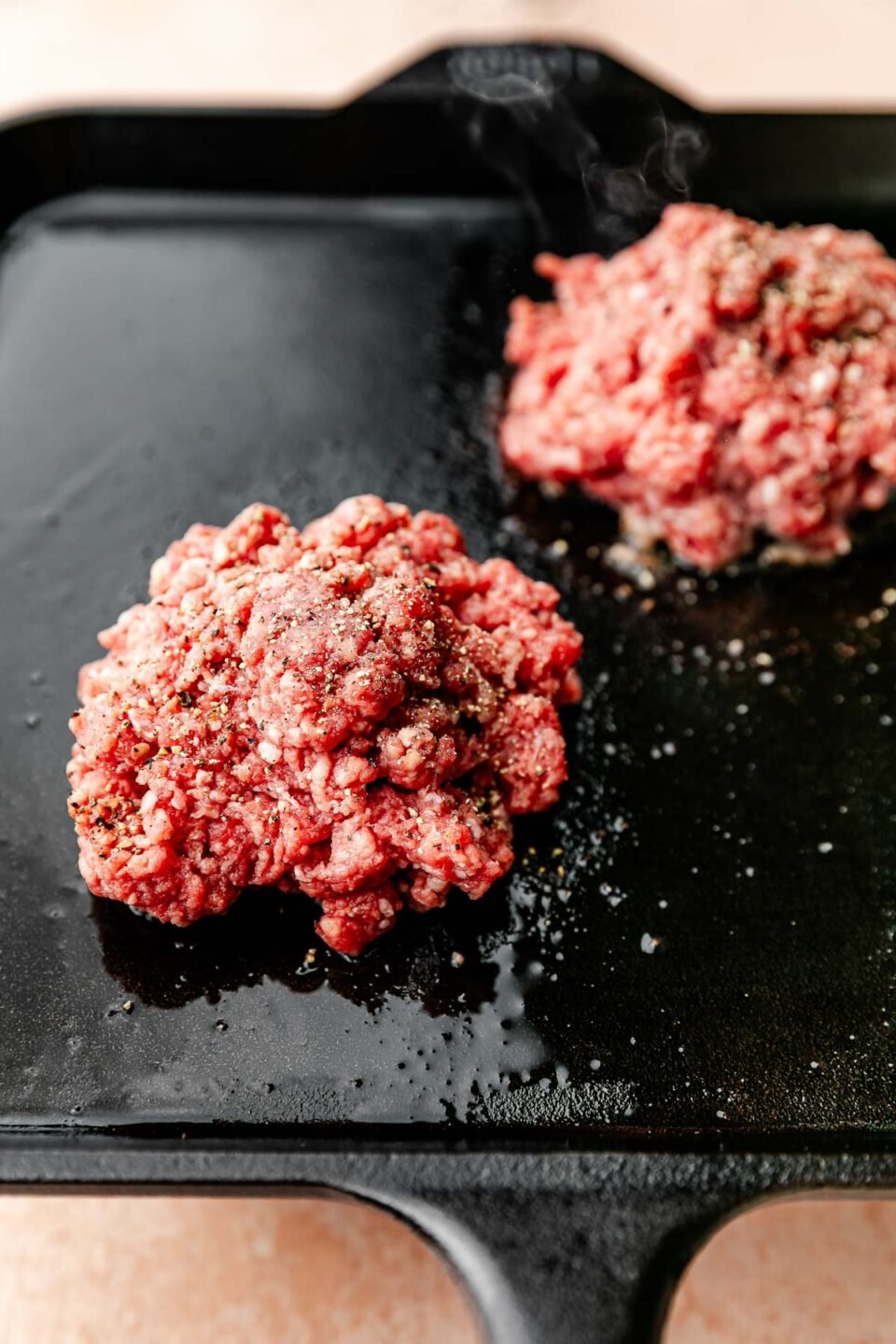 A close-up shot of two raw burger patties atop a black square skillet on a light pink surface.