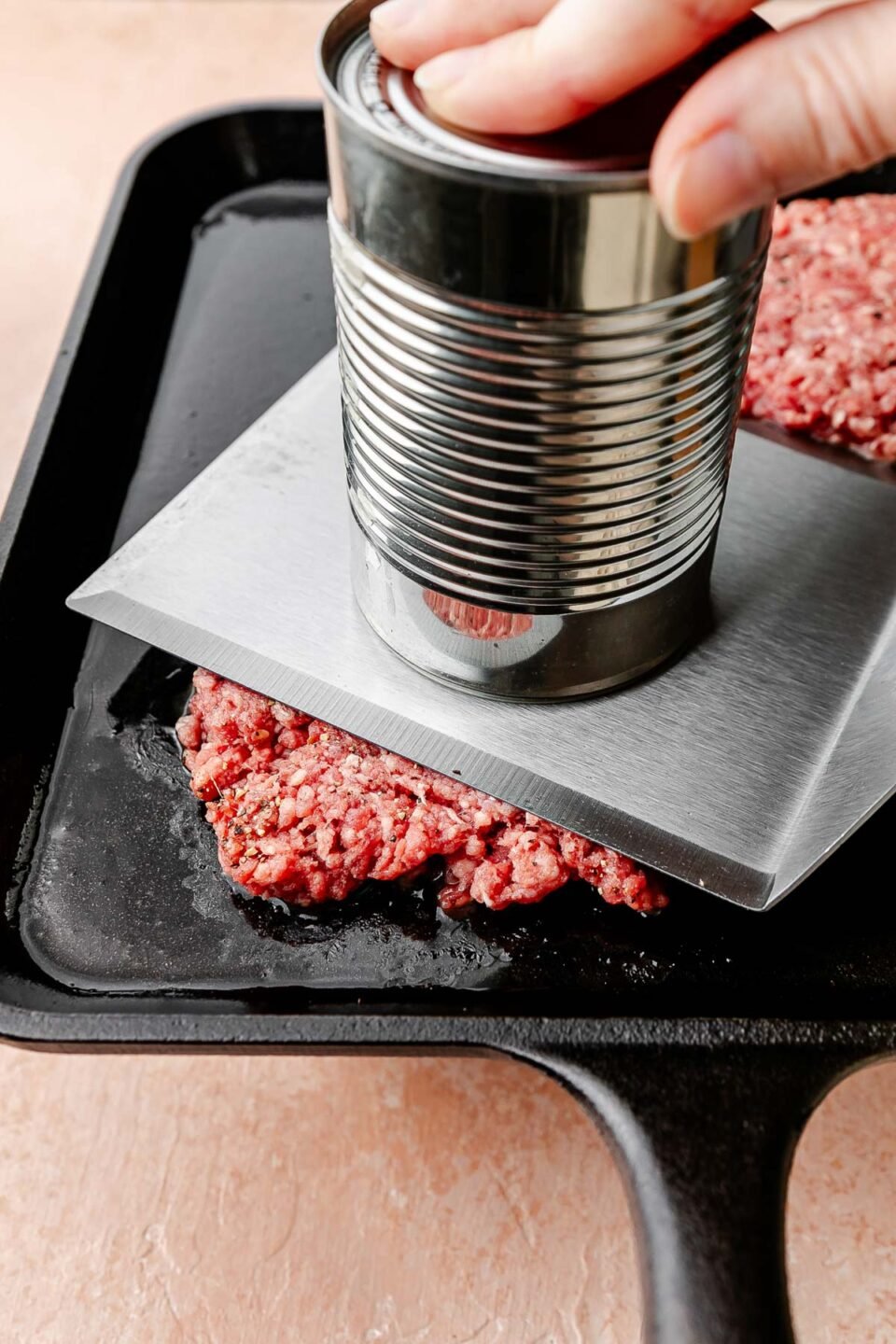 A side-angle shot of a burger patty being smashed on a black skillet with a can and a metal spatula.