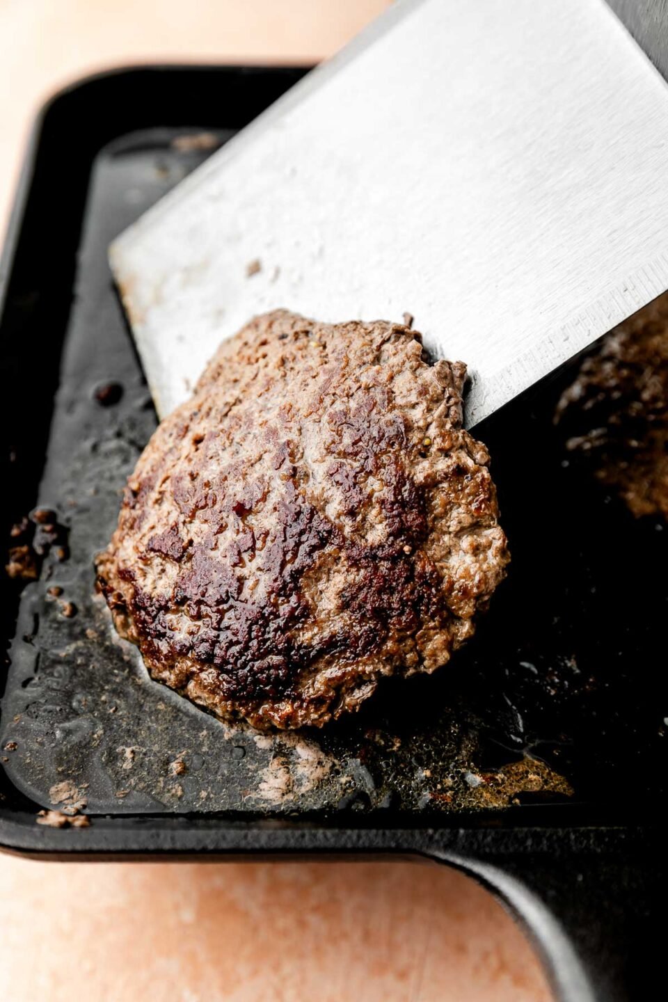 A close-up shot of a smashed burger patty being flipped with a metal spatula on a black skillet atop a light pink surface.