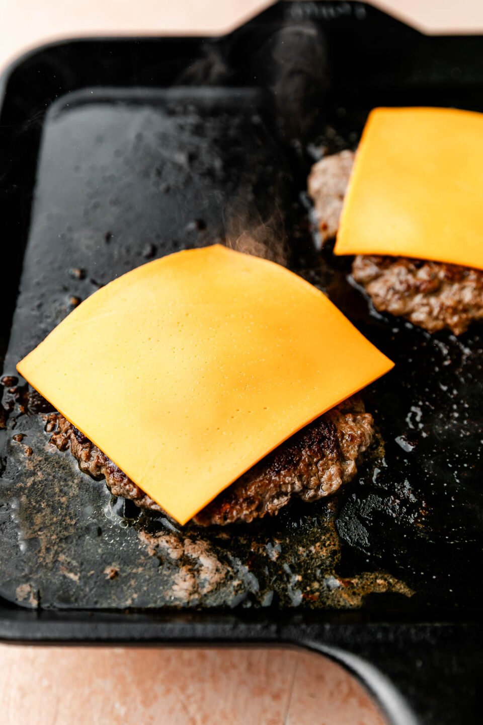 A close-up angled shot of two burger patties topped with cheese slices on a black skillet atop a light pink surface.