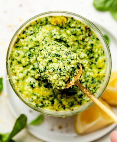 A close-up overhead shot of a spoon holding up basil pesto from a jar. The jar sits on a white plate with lemon wedges atop a white surface.