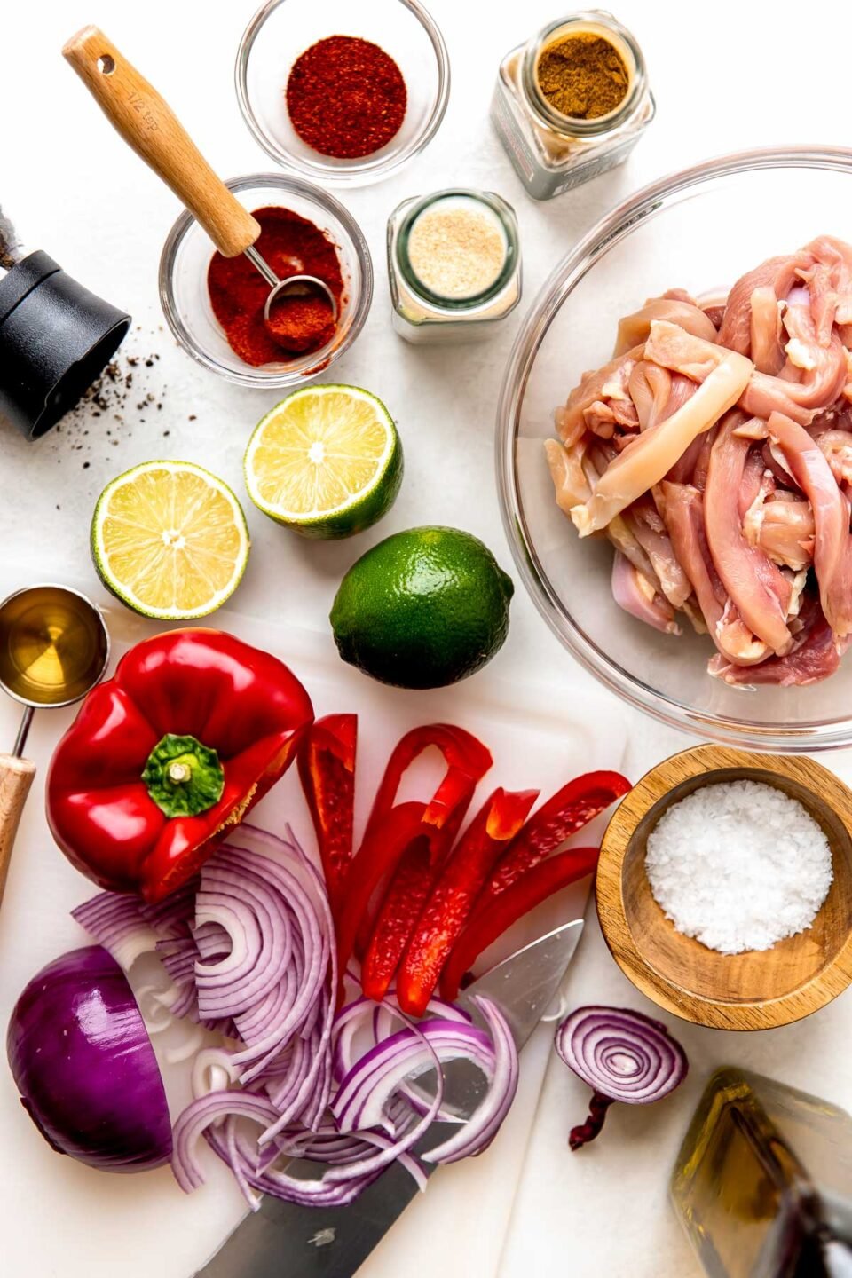 Ingredients are displayed on a white surface: sliced red onion, red pepper, halved lime, jars and bowls of spices, sliced chicken, agave and salt and pepper.