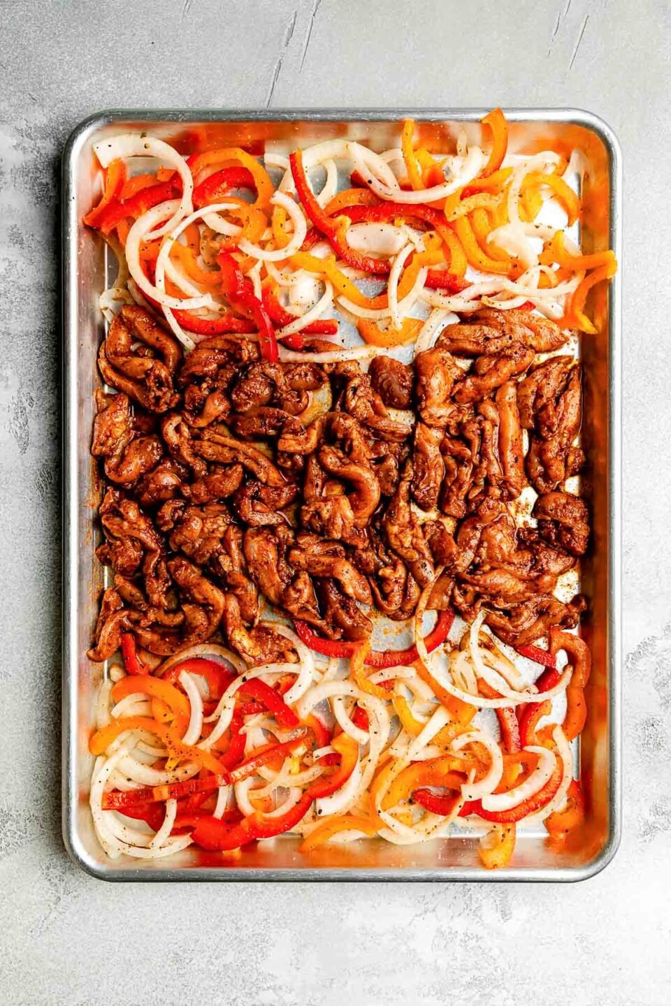 An overhead shot of prepared sheet pan chicken fajita ingredients on a sheet pan atop a light grey surface: seasoned chicken, sliced bell peppers and onions.