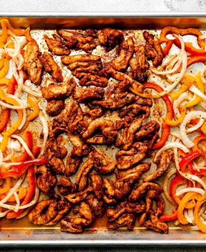 An overhead shot of cooked sheet pan fajitas on a sheet pan atop a light grey surface: seasoned chicken, sliced bell peppers and onions.