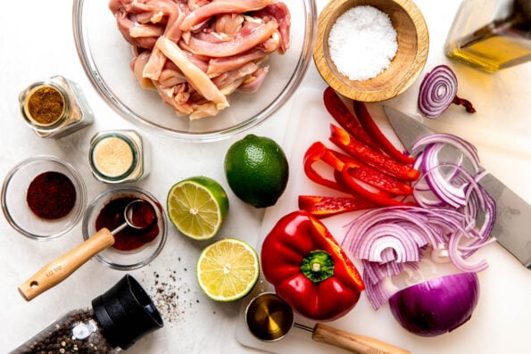 Ingredients are displayed on a white surface: sliced red onion, red pepper, halved lime, jars and bowls of spices, sliced chicken, agave and salt and pepper.