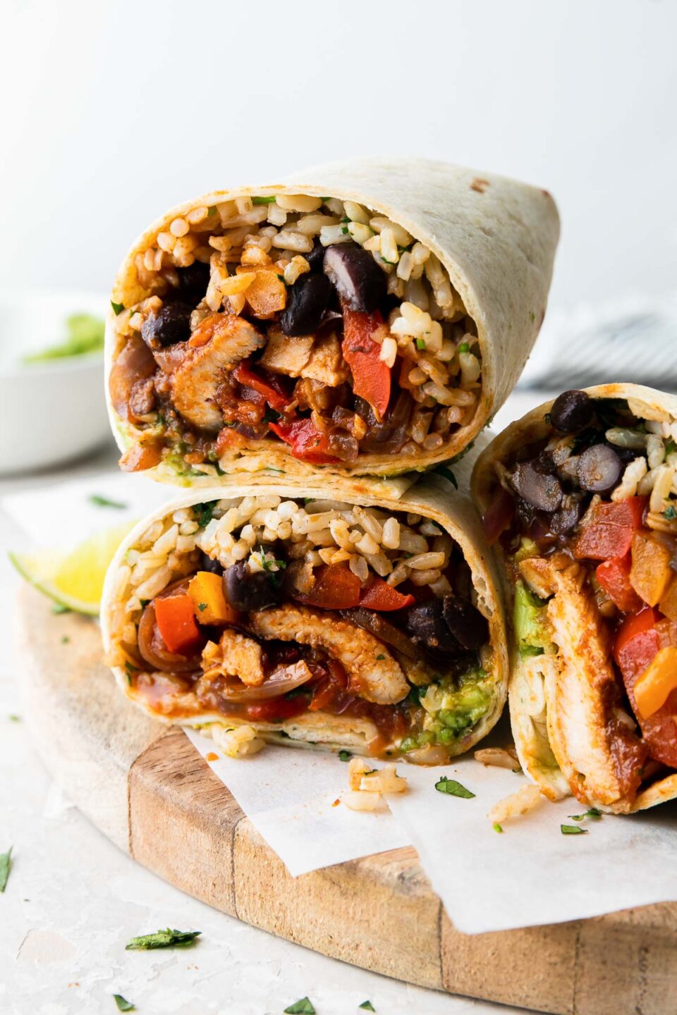 A side angle shot of three stacked burritos stuffed with baked fajita chicken, rice, peppers, black beans and guacamole on parchment paper on a wooden tray atop a white surface.