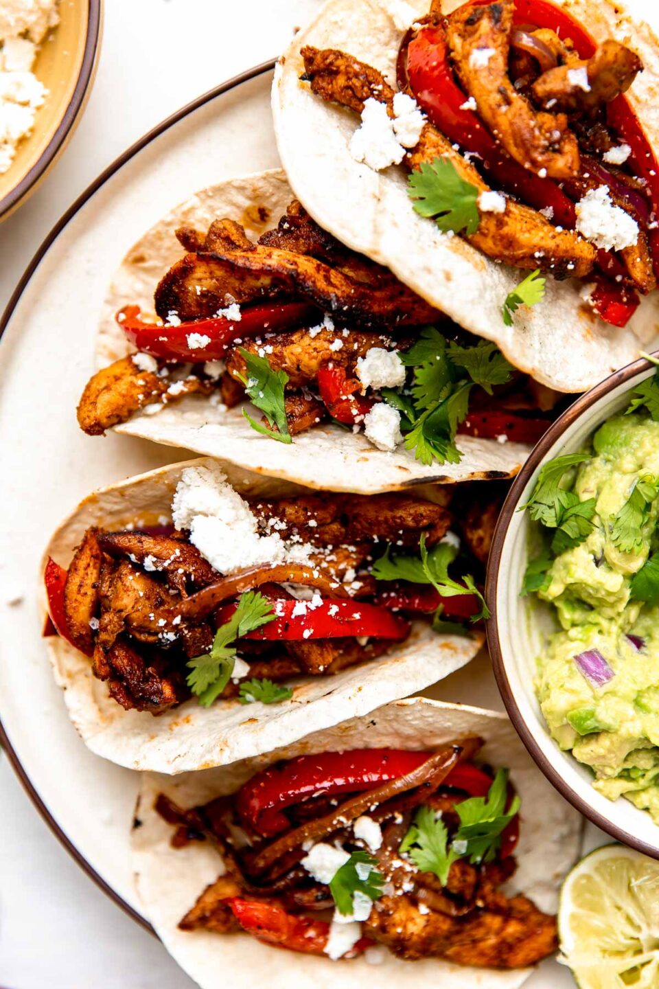 An overhead shot of four sheet pan chicken fajita tacos on a white plate alongside a bowl of guacamole. The plate sits on a white surface.