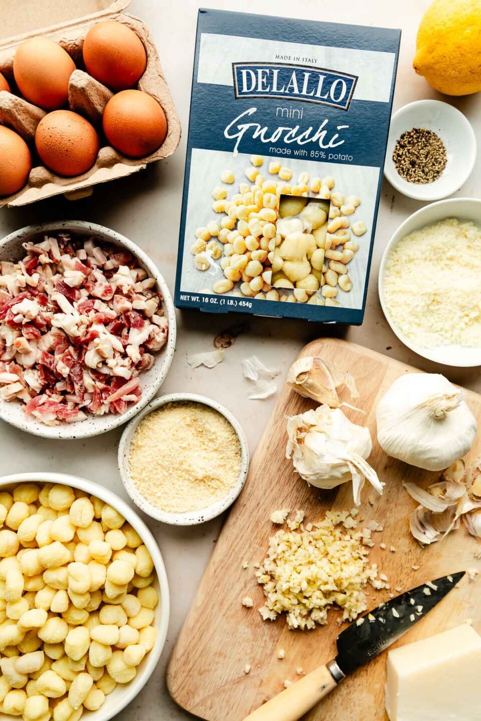 Ingredients are displayed on an off-white surface: mini gnocchi, chopped garlic, pancetta, eggs, parmesan, lemon, black pepper, and pecorino romano.