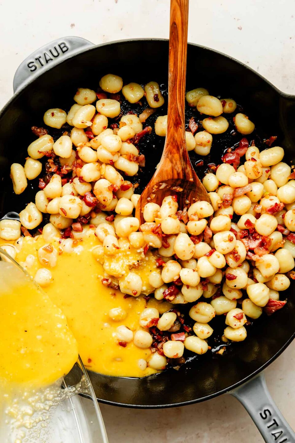 An overhead photo of sauce being poured over gnocchi and pancetta in a black cast-iron skillet atop a beige surface.