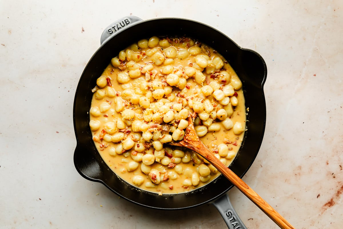 An overhead shot of gnocchi carbonara with a wooden spoon in a black cast iron skillet atop a beige surface.