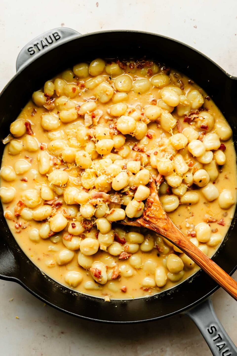 An overhead shot of gnocchi being mixed with sauce with a wooden spoon in a black cast iron skillet atop a beige surface.