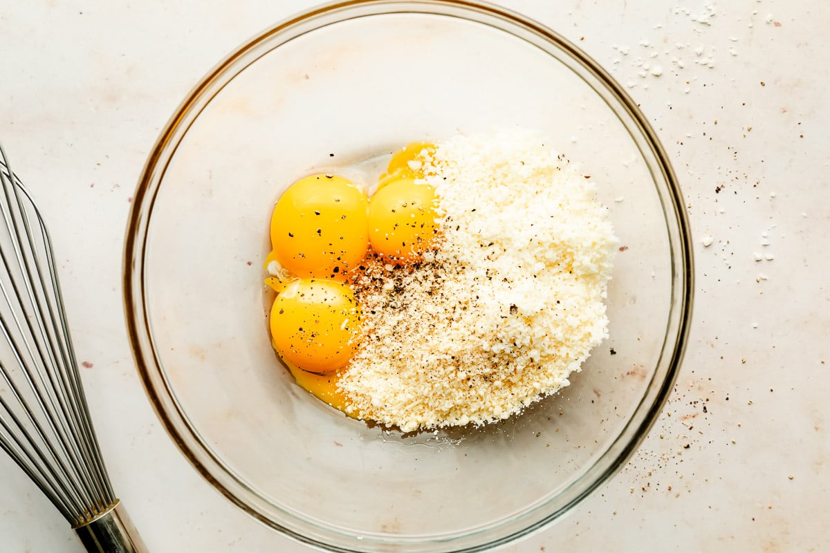 An overhead shot of eggs, cheese, and pepper in a glass bowl atop an off-white surface. A whisk sits beside the bowl.