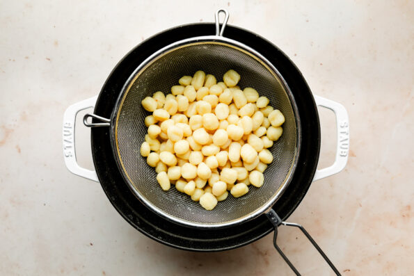 An overhead shot of cooked gnocchi in a colander over a Dutch oven sitting atop a beige surface.