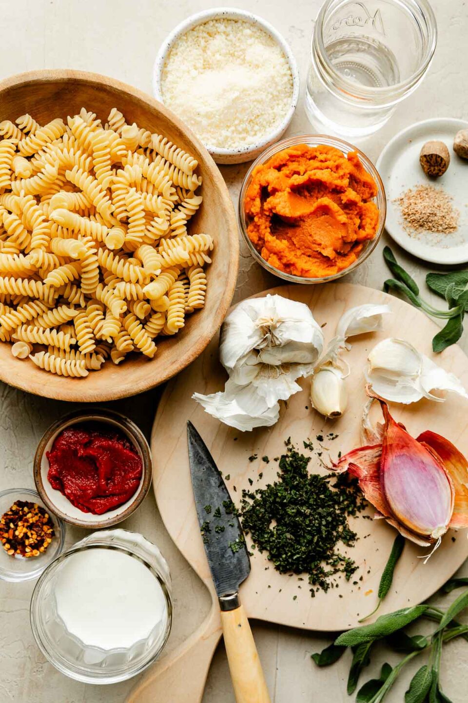An overhead shot of ingredients displayed on a wooden board and an off-white surface: dry fusilli noodles, shallot, garlic, sage, pumpkin puree, nutmeg, red pepper flakes, tomato paste, vodka, parmesan and cream.