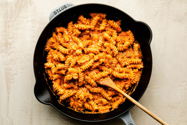 An overhead shot of pumpkin pasta alla vodka with a wooden spoon in a black cast-iron skillet atop an off-white surface.