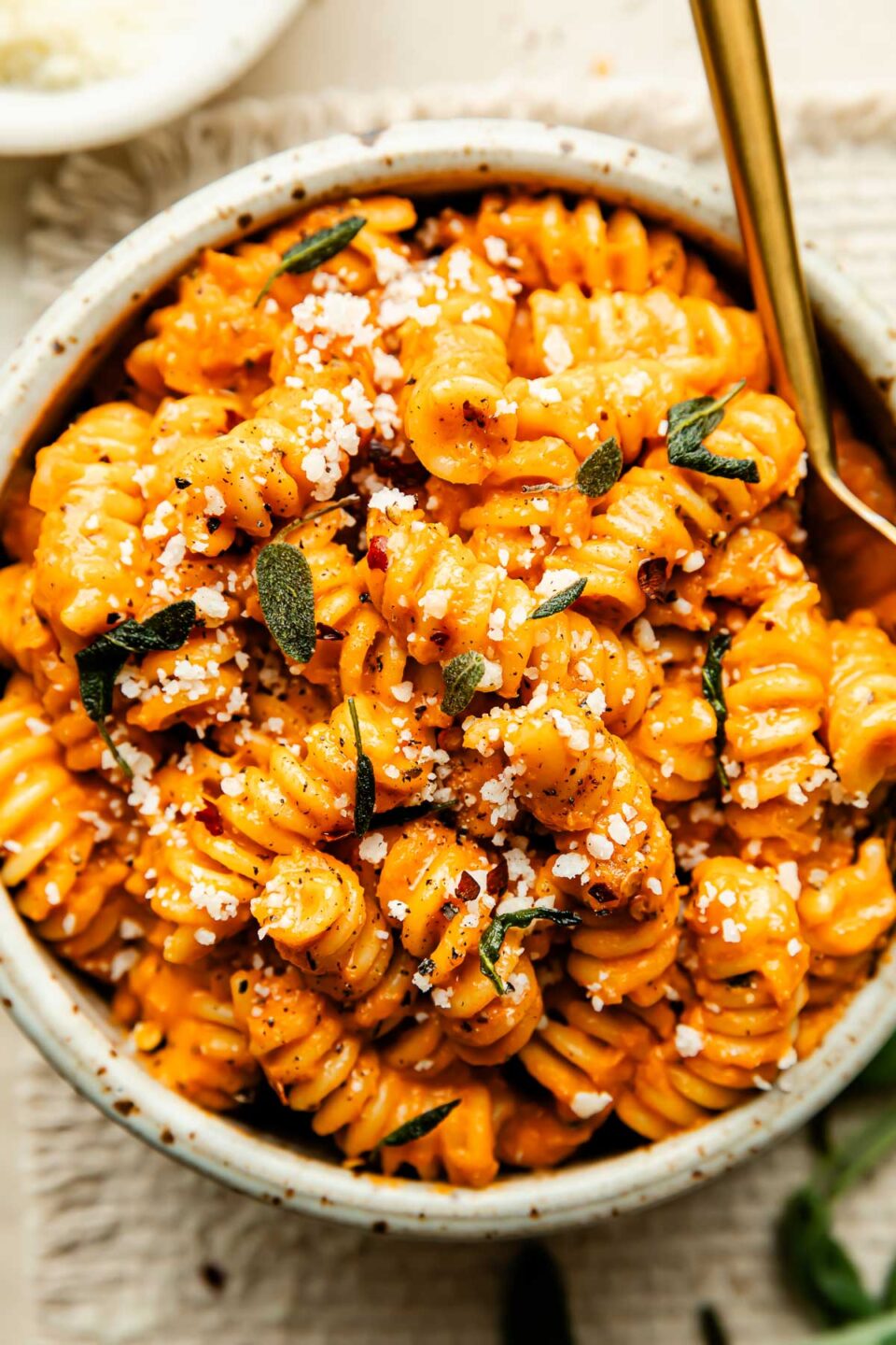 An overhead shot of an off-white stoneware bowl of pumpkin pasta alla vodka topped with grated parmesan and sage leaves atop a woven cream placemat.