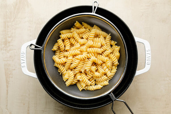 An overhead shot of cooked fusilli noodles in a colander over a cocotte atop an off-white surface.