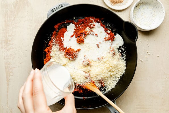 An overhead shot of cream being poured over sauce base in a black cast-iron skillet atop an off-white surface.