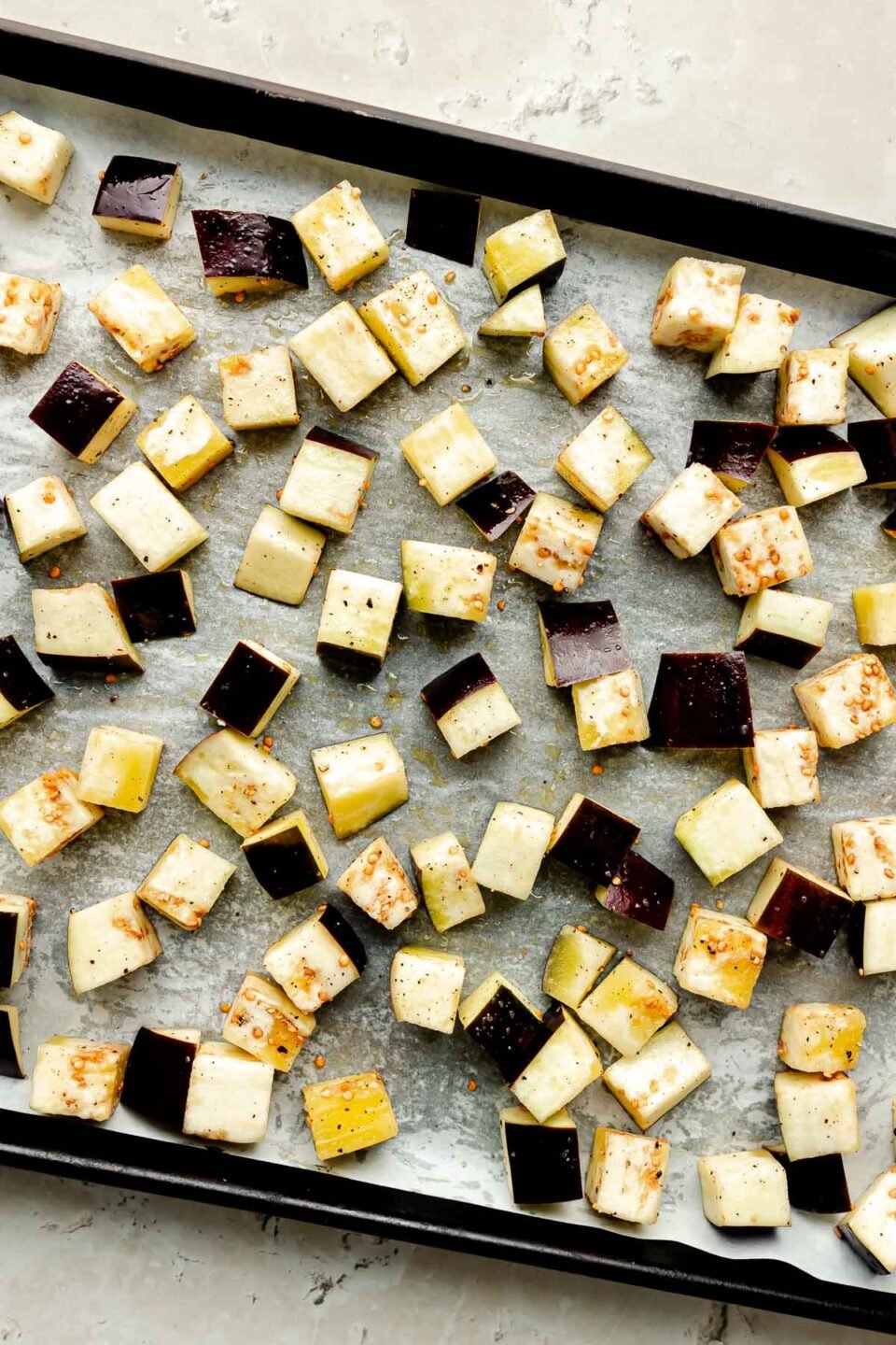 An overhead shot of a sheet pan of diced eggplant on parchment paper atop a beige surface.