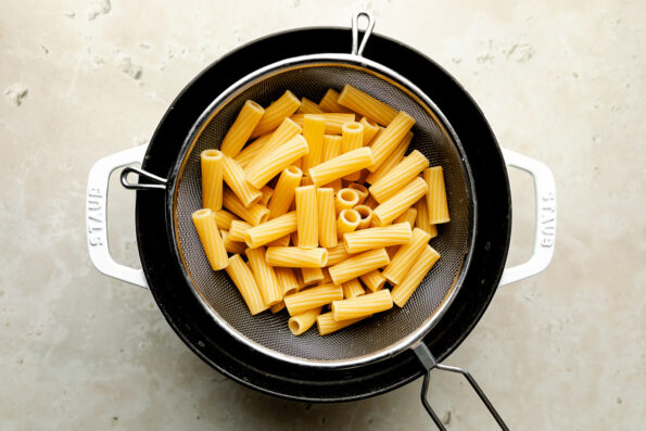 An overhead shot of cooked tortiglioni noodles in a colander over a pot atop a beige surface.