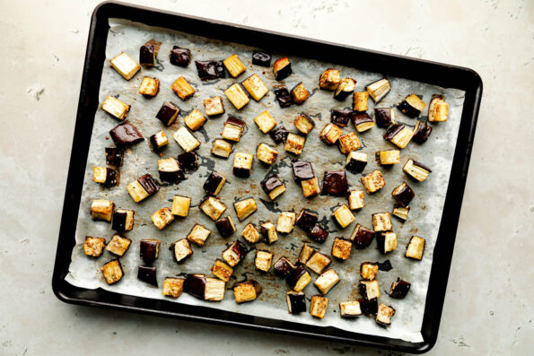 An overhead shot of roasted eggplant on parchment paper on a sheet pan. The sheet pan sits atop a beige surface.